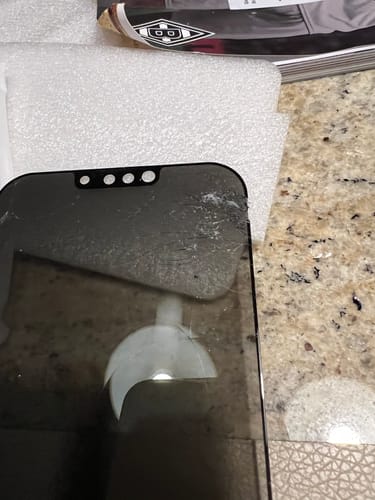 Nahaufnahme der beschädigten 3 in 1 Schutzfolie für ein iPhone, die auf einer Schaumstoffverpackung liegt.