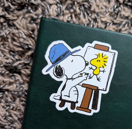 Customer's Snoopy sticker of Snoopy in a blue hat painting Woodstock, placed on a green journal.