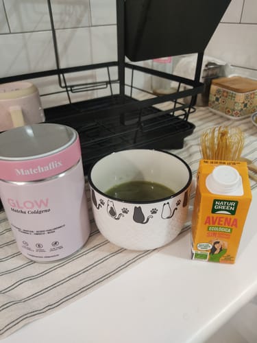 Cliente muestra el producto Matchaflix Glow, una taza de matcha y un cartón de leche de avena en una encimera de cocina.