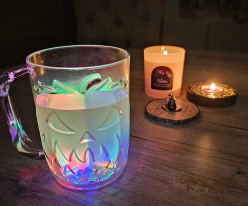 Rezensent zeigt den Spooky LED-Becher in Kürbisform, der auf einem Tisch neben Kerzen bunt leuchtet.