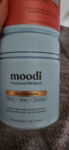 Reviewer holds a tub of Moodi PM Blend, showing the 'Choc Hazelnut' flavor.