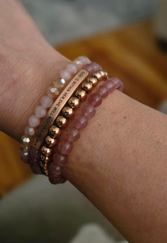 A reviewer's wrist wearing the stacked 'GOD IS WITHIN HER' bracelet bundle, highlighting the engraved bar and beaded bracelets.