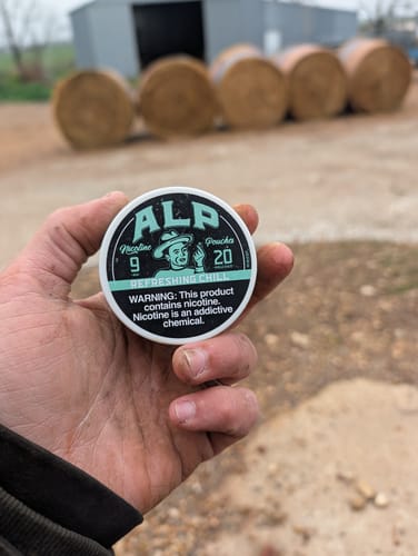 Customer's hand holding a can of Refreshing Chill Nicotine Pouches outdoors with hay bales in the background.