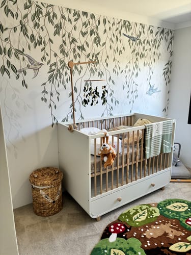 Reviewer's Under the Willow Wallpaper Mural on the wall behind a white crib in a lovely, decorated nursery.