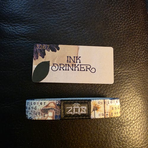 Customer's Inkdrinker Bracelet with its collector's card on a black surface. The bracelet features a detailed collage design.