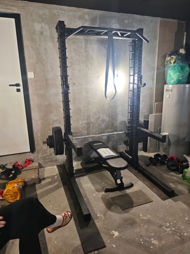 Rezensent zeigt das R7 Saber Squat Rack mit Multipresse, aufgebaut in einer Garage mit Hantelbank und Langhantel.