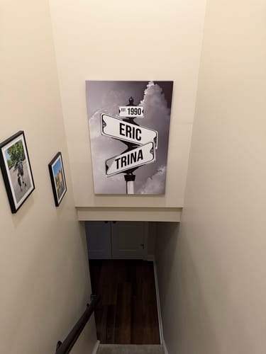 Customer's personalized street sign canvas with 'Eric', 'Trina', and '1990' hanging on a wall above a staircase.