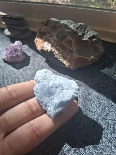 Reviewer holding a rough, light blue calcite crystal in their palm with other crystals in the background.