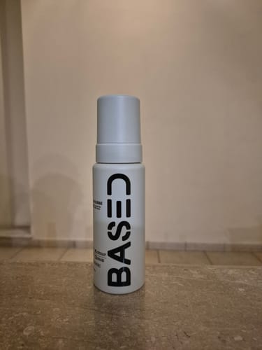 Reviewer's white bottle of Based Curl Mousse with a grey cap standing on a stone surface.