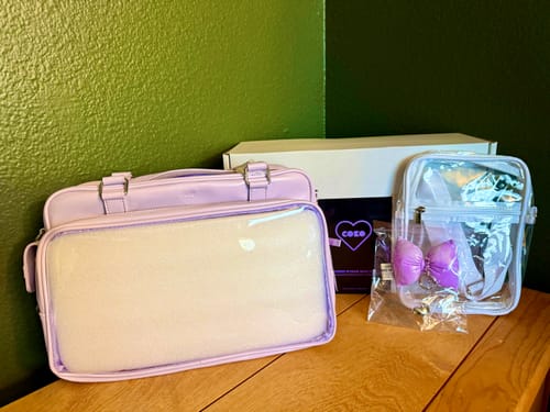 Customer's lilac COKO STAGE BAG with its box, a smaller clear pouch, and a bow keychain on a wooden surface.
