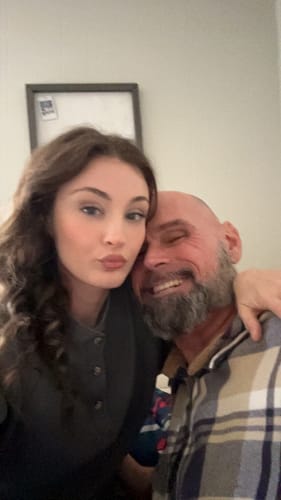 Close-up selfie of a happy reviewer, a man with a beard, being embraced by a woman.