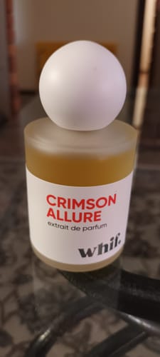 A reviewer's bottle of Crimson Allure perfume, showing its frosted bottle and round white cap on a dark tabletop.