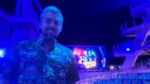 Reviewer smiling in their 'Flux Overload' shirt at an outdoor venue lit by blue lights at night.