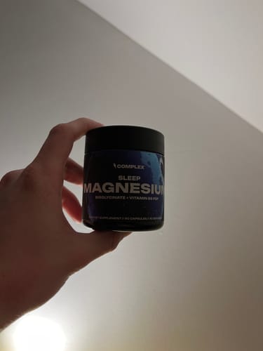 Customer's hand holding a dark tub of Complex Sleep Magnesium supplement indoors.