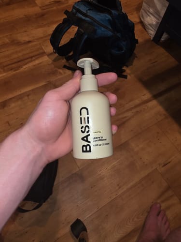 A customer holds the Based Bodyworks Leave-in Conditioner in a beige pump bottle against a wooden floor background.