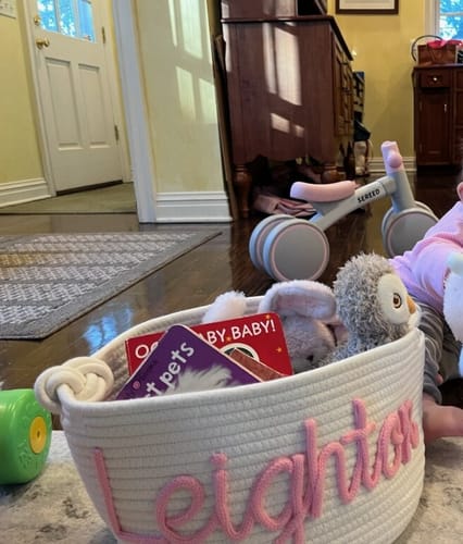 Customer's white personalized knitted basket with 'Leighton' in pink yarn, filled with toys and books on a floor.