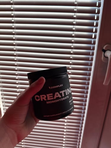 A customer's hand holding a black tub of Creatine Monohydrate in front of a window with blinds.