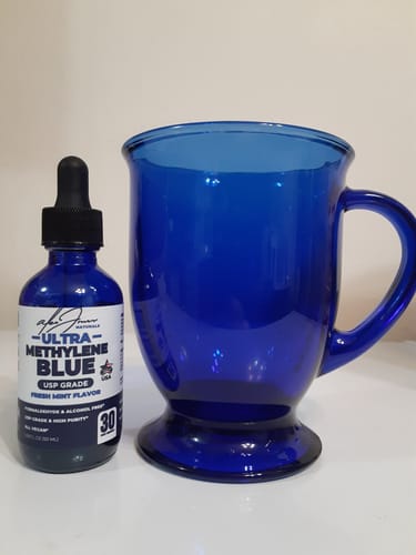 Customer's bottle of Ultra Methylene Blue next to a large blue glass mug on a white surface.