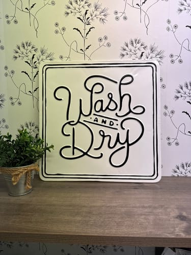 Reviewer's white Wash And Dry Tin Sign with black script text sitting on a wooden laundry room shelf.