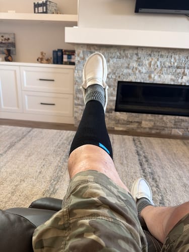 A first-person view of a customer wearing a black Performance Eucalyptus Compression Sock while relaxing indoors.