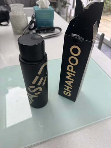 Customer's black shampoo bottle standing next to its open black box with gold lettering on a glass table.