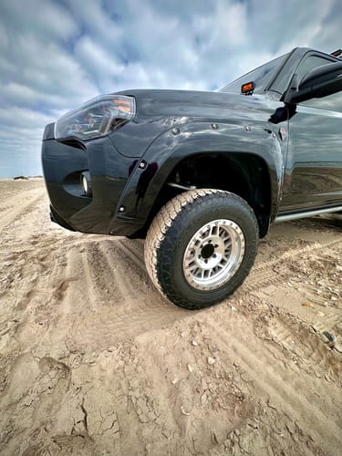 Customer's Forged Aluminum Upper Control Arms installed in the wheel well of a black Toyota 4Runner on sand.
