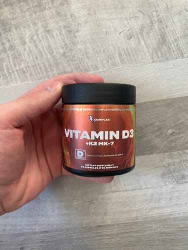 Customer's hand holding a black jar of Vitamin D3+K2 MK-7 supplement against a wood-grain surface.