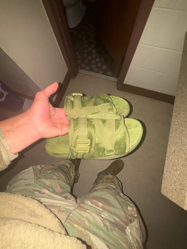A customer holds up their new olive green Mystery DracoSlides, which have a plastic buckle and match their camouflage pants.