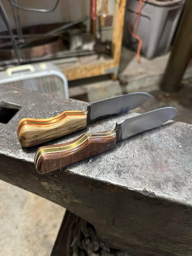 Customer's two finished knives with custom wood handles resting on a metal anvil in a workshop.