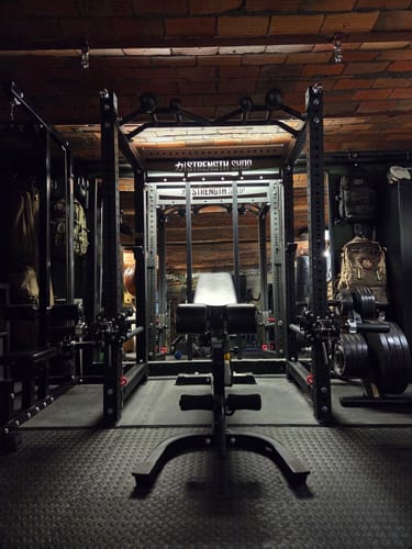 Customer's Riot MRR 75 Compact Power Rack with a bench, viewed from the front in a home gym with a low brick ceiling.