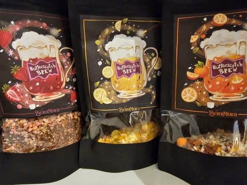 Kunde zeigt die drei fruchtigen Sorten des Butterscotch Brew Tees – Erdbeere, Zitrone und Orange – in ihren schwarzen Verpackungen.