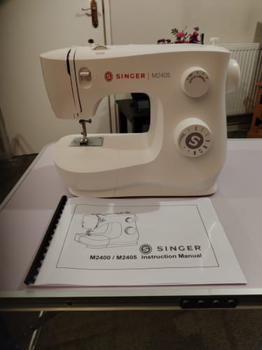 Customer's printed Singer sewing machine manual displayed on a table in front of a white Singer M2405 sewing machine.