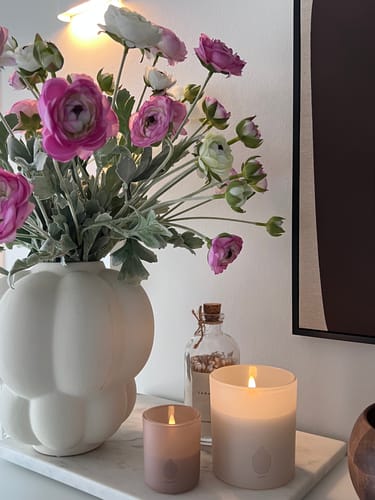 Recensent toont het Ranonkel Arrangement in een witte, decoratieve vaas naast twee brandende kaarsen.