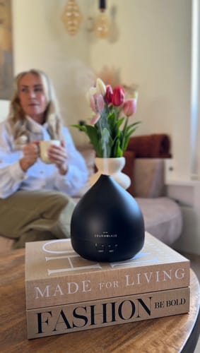 Recensent toont de matte zwarte Unity 2.0 diffuser werkend op een stapel boeken in een sfeervolle woonkamer.