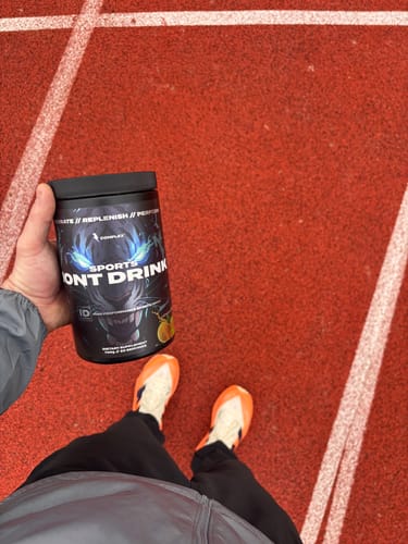 Reviewer holding a black tub of Sports Iont Drink while standing on a red running track.