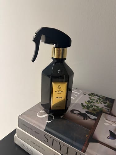 Reviewer's ANASS room freshener, a black spray bottle with a gold label, displayed on a stack of books.