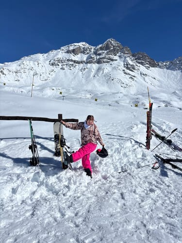 Reviewer wearing pink baggy snow pants on a snowy mountain, posing with a snowboard.
