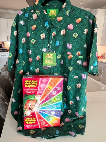 Reviewer's green Lucky Charms shirt on a hanger, displayed with a box of Lucky Charms cereal in front.