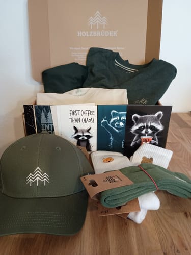 Rezensent zeigt die geöffnete Holzbrüder Wunderbox mit Kappe, T-Shirts, Socken und Postkarten.