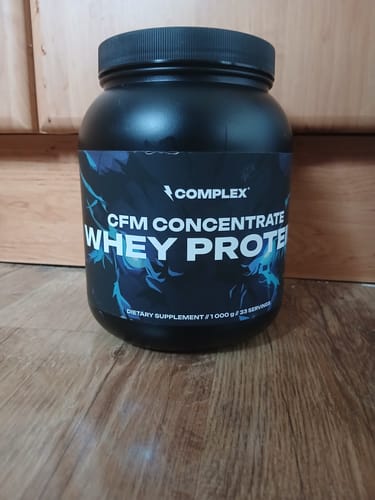 Customer's black tub of CFM Whey Protein on a wood floor in front of a cabinet.