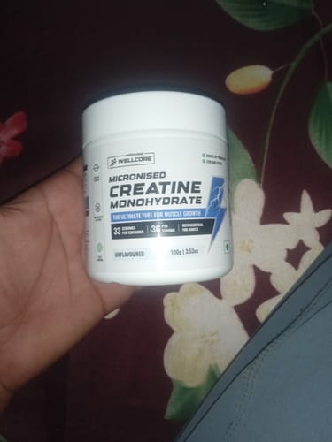 A customer's hand holding a white tub of Wellcore Micronised Creatine Monohydrate against a dark background.