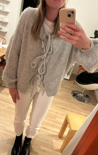 Kundin trägt den gemütlichen Oversized Cardigan mit Schleifen in Grau zu einer weißen Hose.