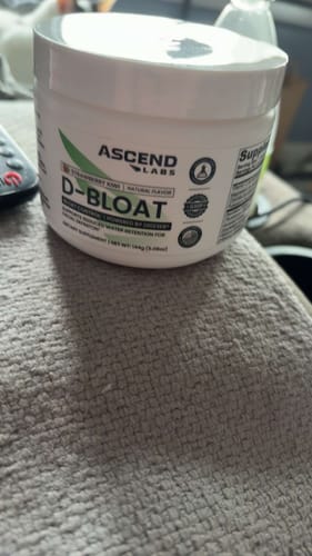 Customer's white tub of D-BLOAT supplement resting on a grey textured surface.