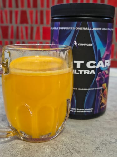 Customer's glass mug of bright orange Joint Care Ultra drink sitting next to the product container.