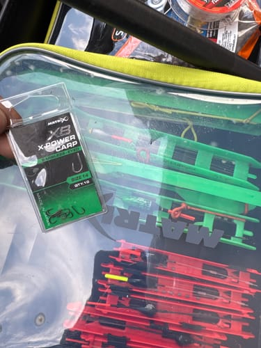 Recensent houdt de verpakking X8 X-Power Carp-haken vast, met op de achtergrond een vistas vol vistuig.