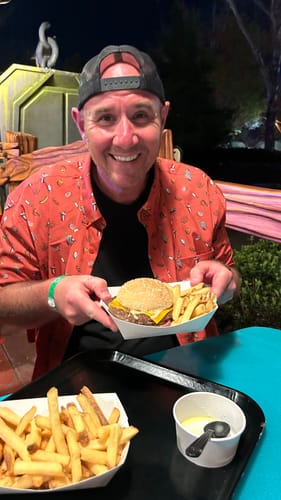 Reviewer smiling while wearing the red Park Bites shirt and holding a burger and fries at a theme park.