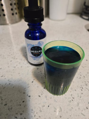 Reviewer's bottle of Meraki Blu Methylene Blue next to a small cup filled with the dark blue liquid on a counter.
