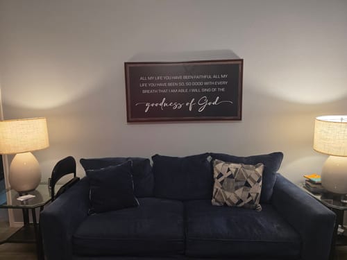 Customer's Goodness Of God Faith Sign II hanging on a wall above a blue sofa in their living room.