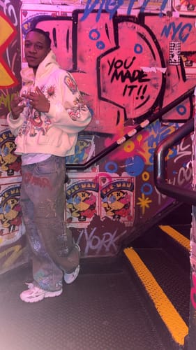 Reviewer posing in a cream graphic hoodie and wide jeans against a graffiti wall.