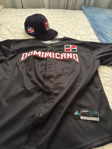 Customer's black Dominican Republic Heritage baseball jersey and matching cap laid out on a bed, showing the 'Dominicano' text.